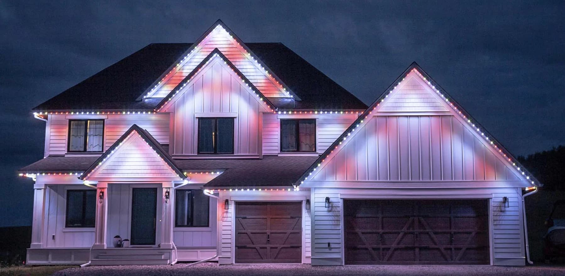 A two-story home at night with permanent lighting in white and soft pink along every gable and eave