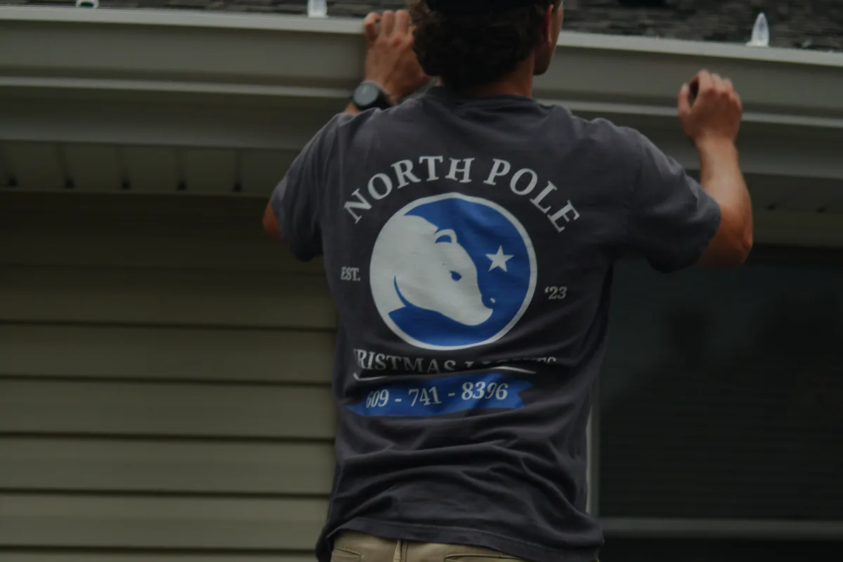 A North Pole installer reaching up to run a strand along the soffit, the bear mark across the back of his shirt